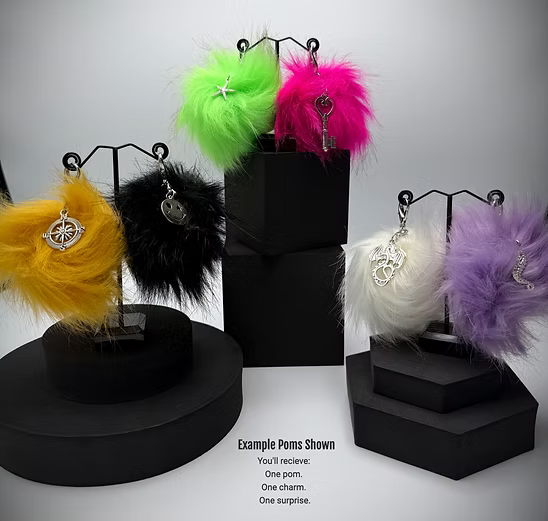 Colorful pom-pom keychains with charms on black stands against a white background.