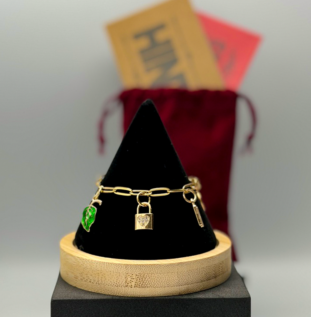 Gold bracelet with charms on a black cone-shaped stand with a blurred red bag in the background.